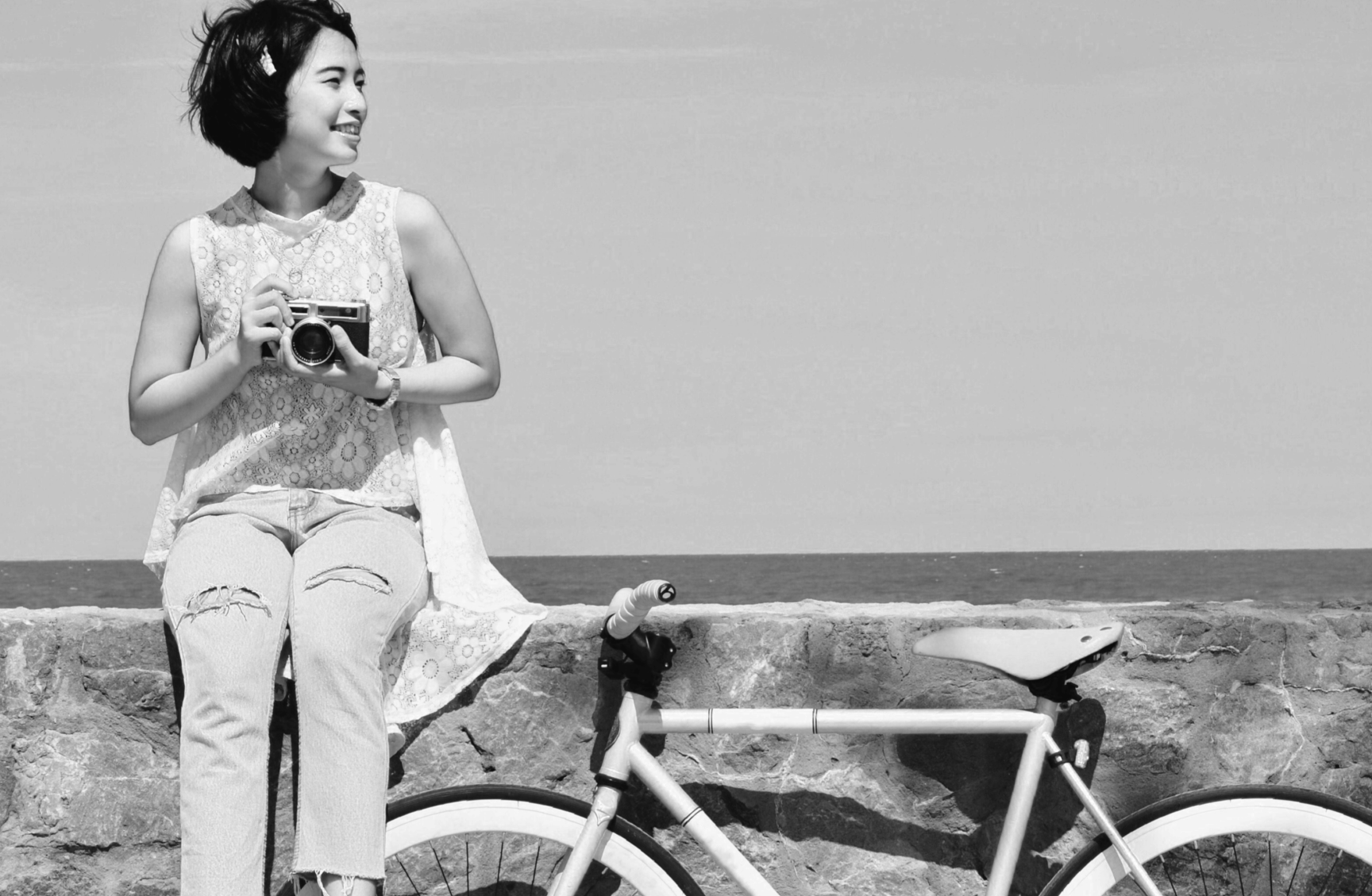 Young Asian woman sitting on stone fence with camera and a bike